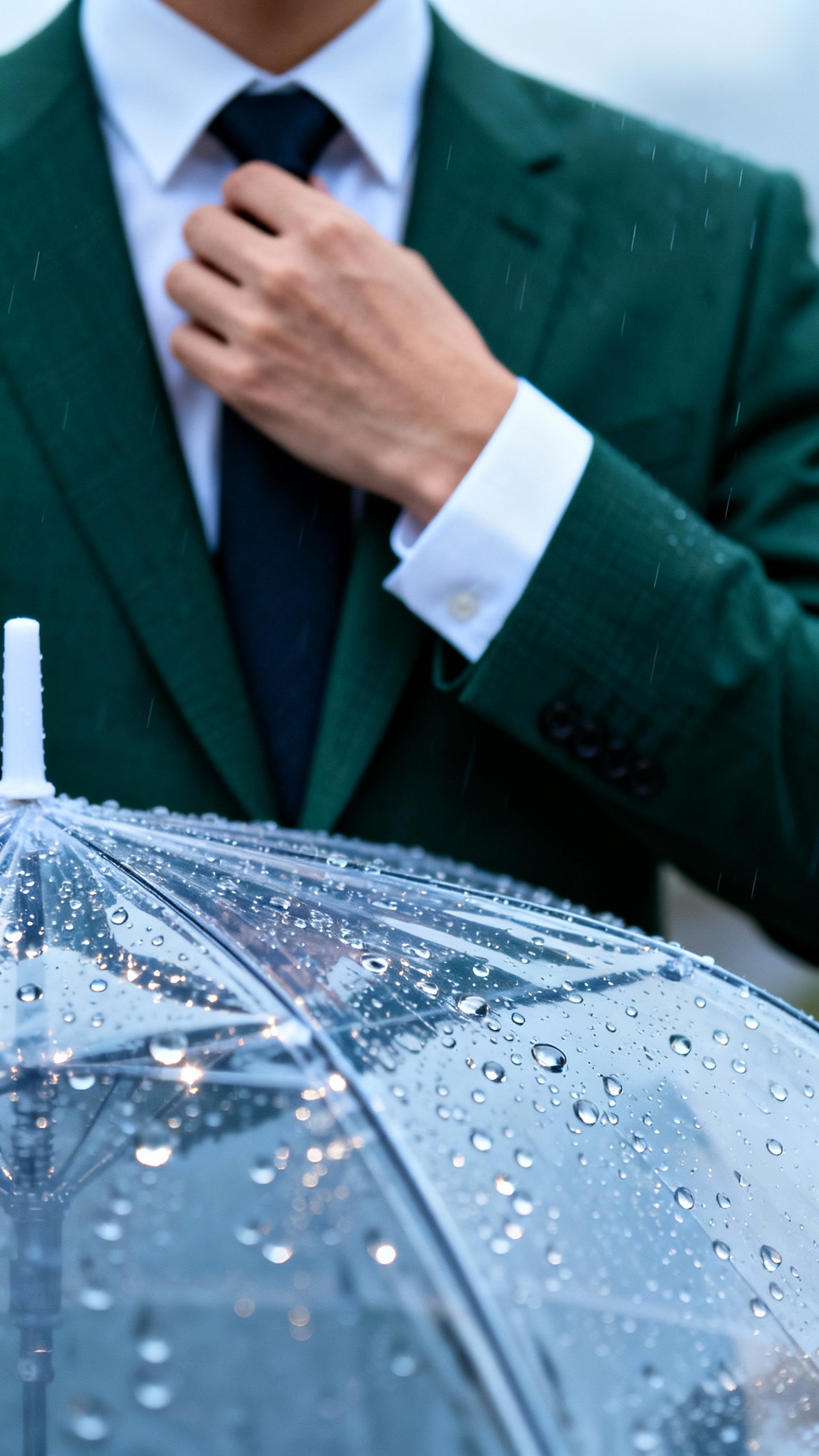 Clear umbrella closeup with raindrops, forest-green suit sleeve adjusting tie