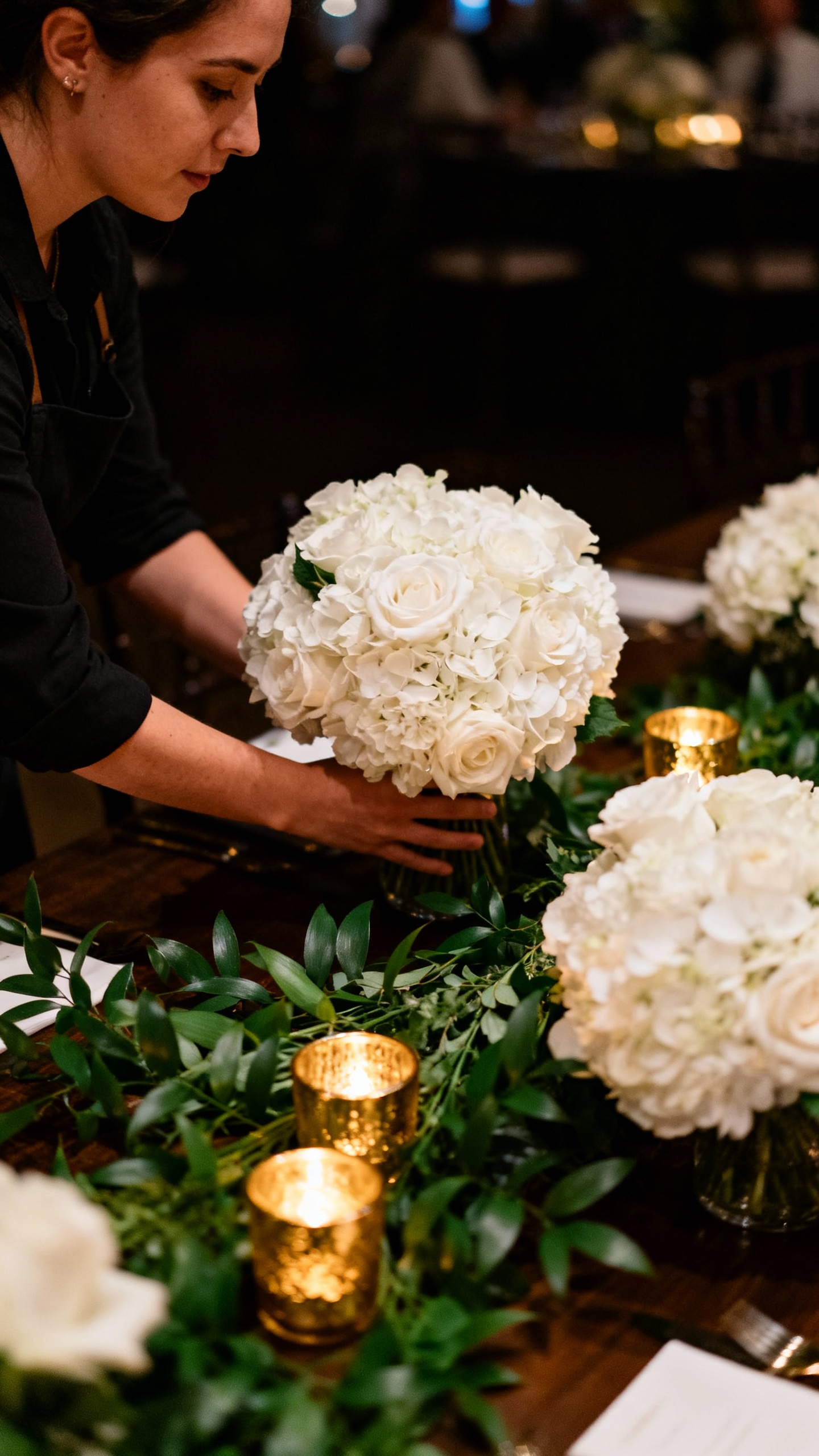 Closeup of florist moving white ceremony arrangements to reception table, lush greenery, gold votive